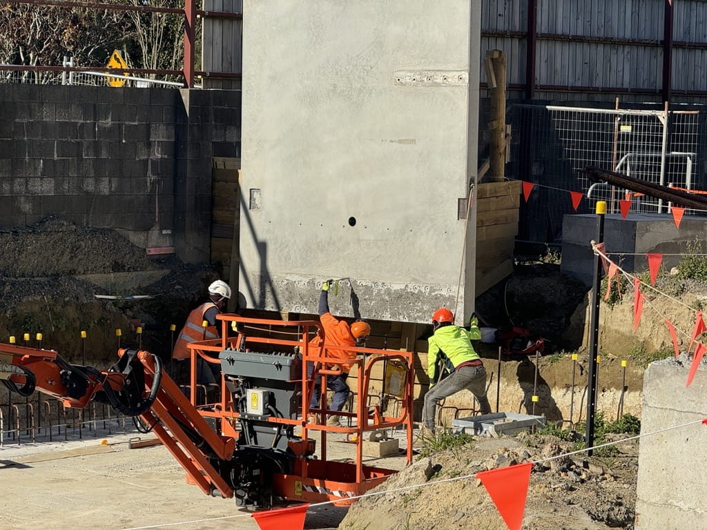 Three workers in PPE helping attach a concrete wall panel being suspended by a crane