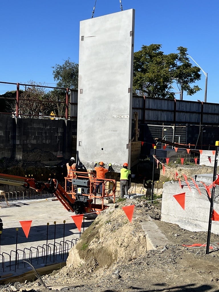 Three workers in PPE helping to put a large concrete panel wall, connected to a crane, into place 